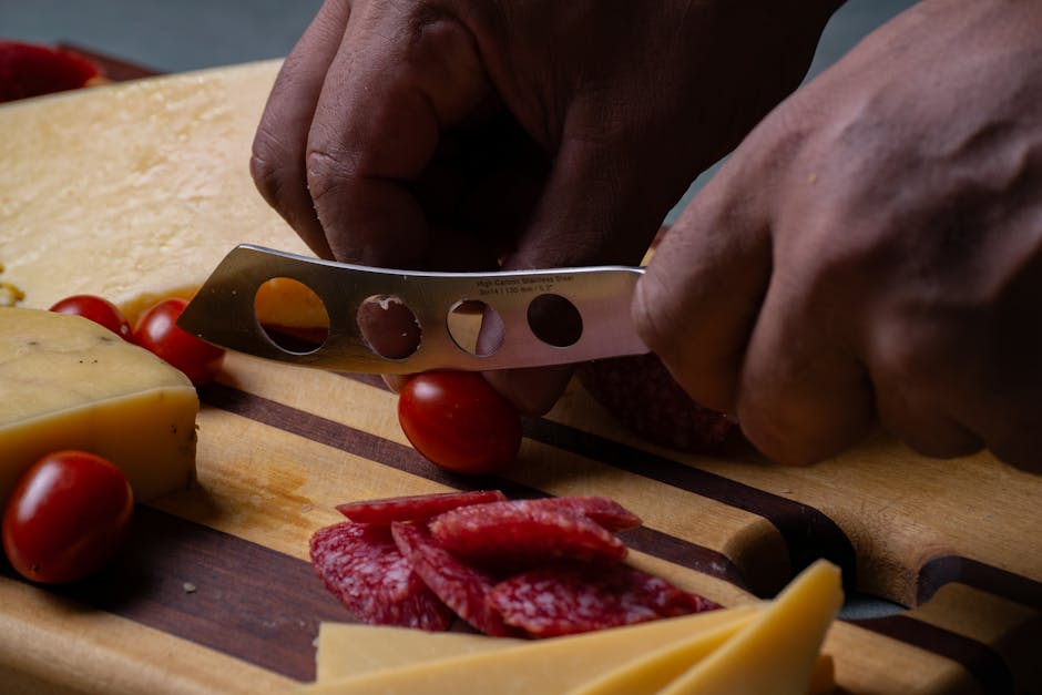 Close-up of hands slicing salami on a meat and cheese platter with knife and cherry tomatoes.